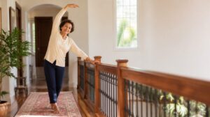 Senior woman in cream cardigan performing balance exercise at a polished wooden handrail in sunny hallway