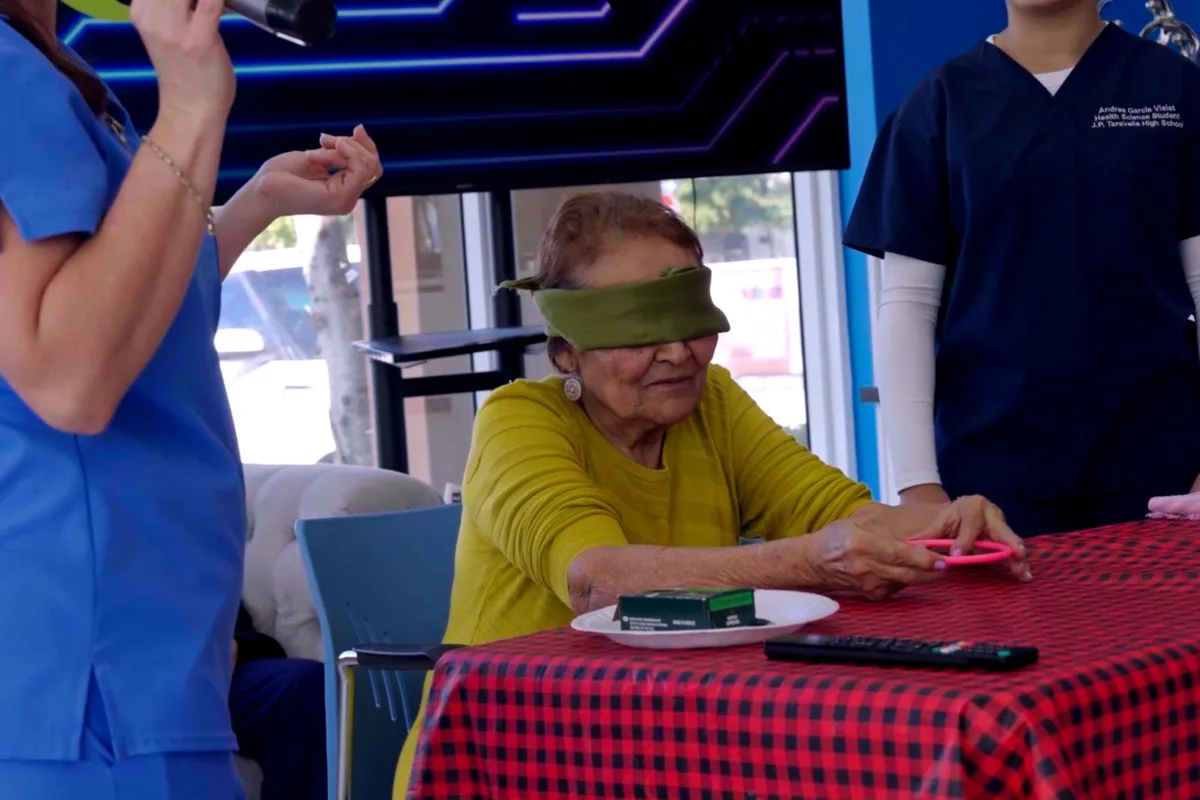 A blindfolded member plays a table game while staff and other members cheer her on