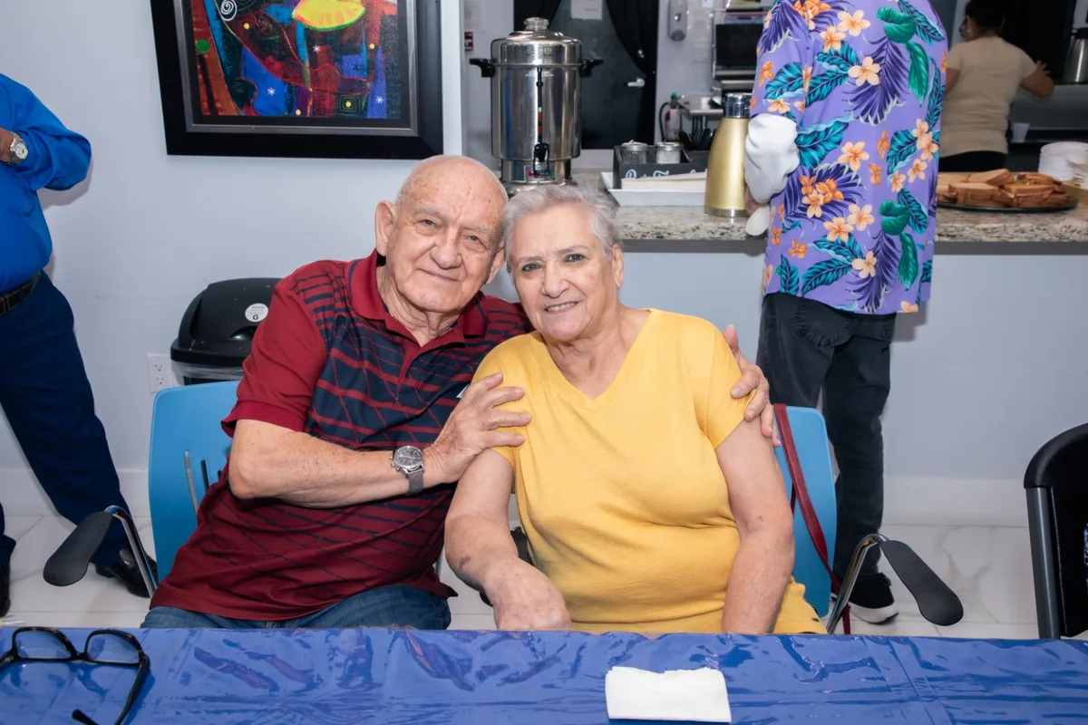 Una pareja de adultos mayores sonríe abrazada, sentados a la mesa durante una actividad en el centro